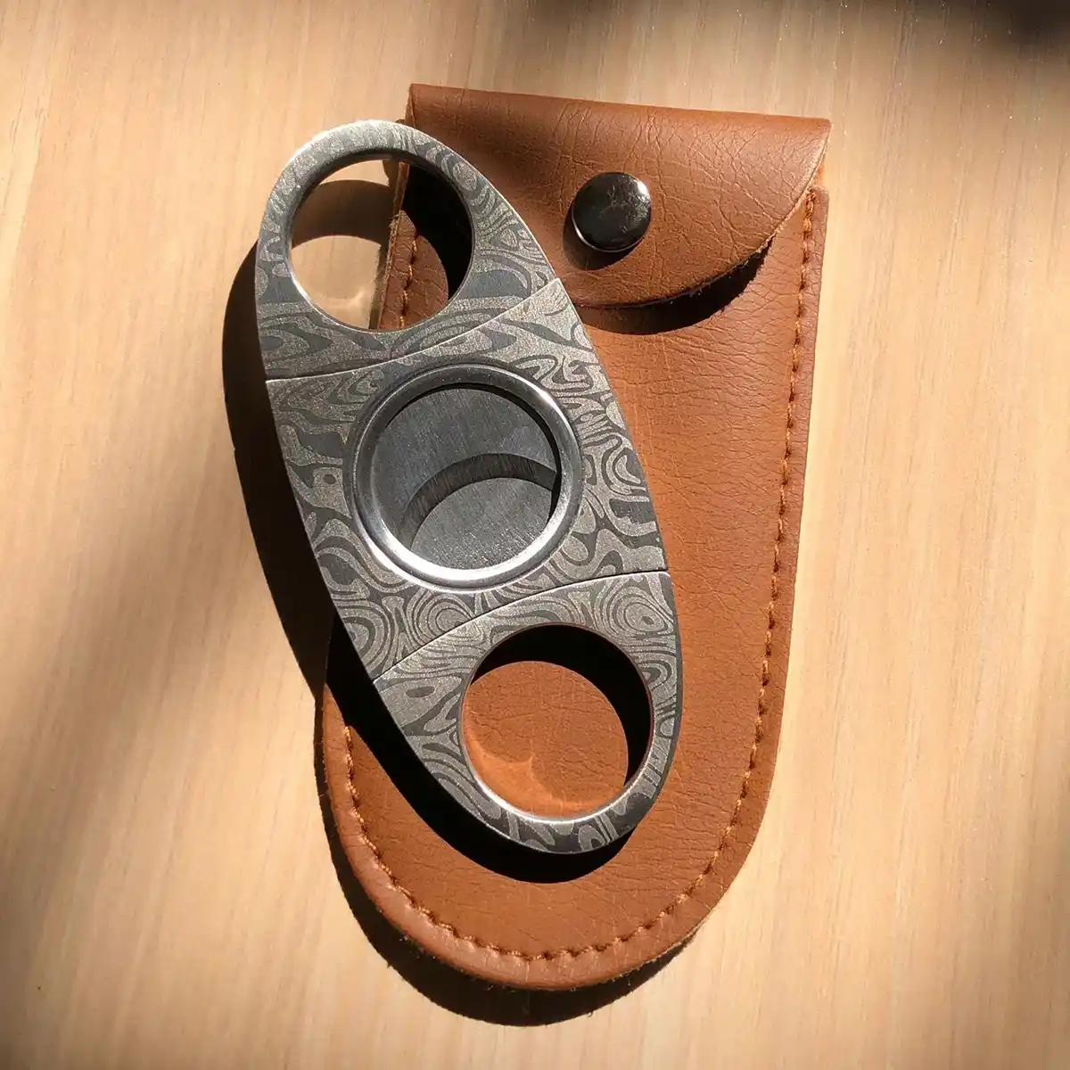 Damascus Cigar Cutter – Double Blade Stainless Steel | ProTravelHumidors 3 A Damascus steel patterned double blade cigar cutter resting on a brown leather pouch ProTravelHumidors