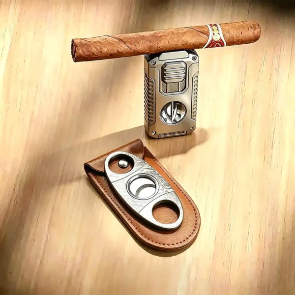 ProTravelHumidors cigar resting on silver torch lighter next to two Damascus steel cigar cutters – one inside a brown leather pouch on a light wooden surface